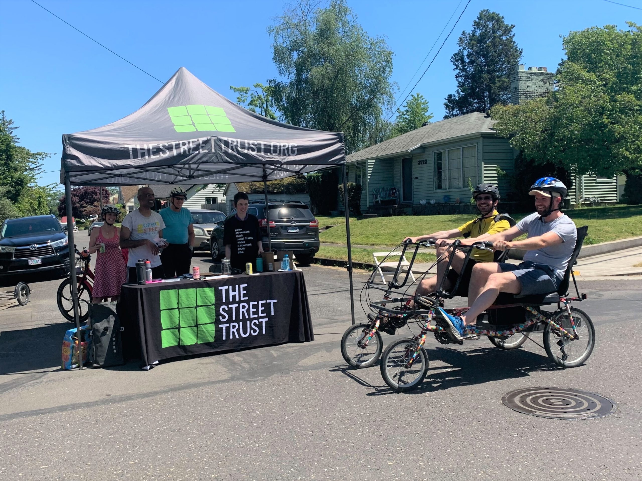 Two people pedal an adaptive quadricycle past The Street Trust's booth