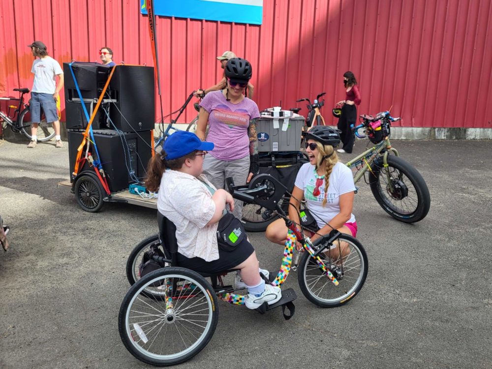 Person sitting on an adaptive handcycle outside of Adaptive BIKETOWN talking to one person who is kneeling and another who is standing. Other people with bikes in background. Bike pulling trailer with sound system in background.