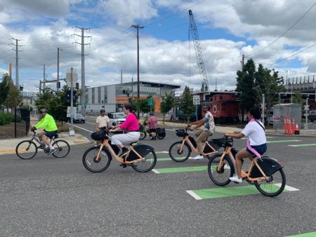 People riding BIKETOWN e-bikes making a left turn while another bike rider in background corks traffic.