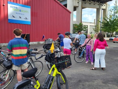 People standing with their bikes socializing outside of a red building with a banner that says Kerr Bikes and Boats Rentals. Some people are dressed in Harry Styles inspired outfits.