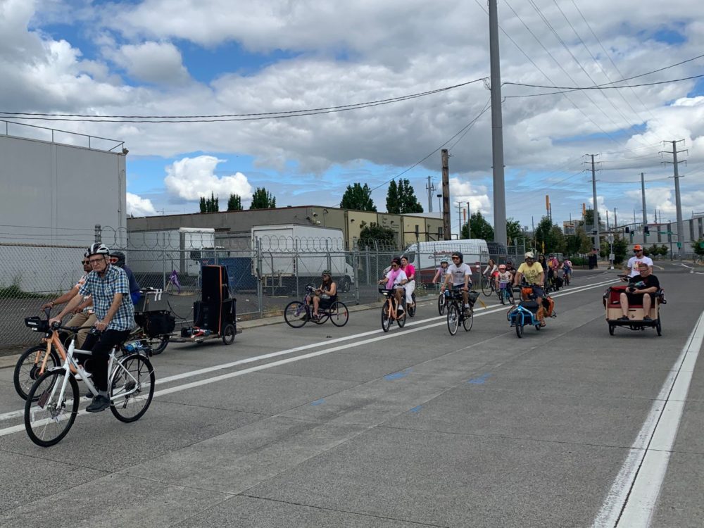 Group of people riding traditional, cargo, and adaptive bikes down empty street in an industrial area.