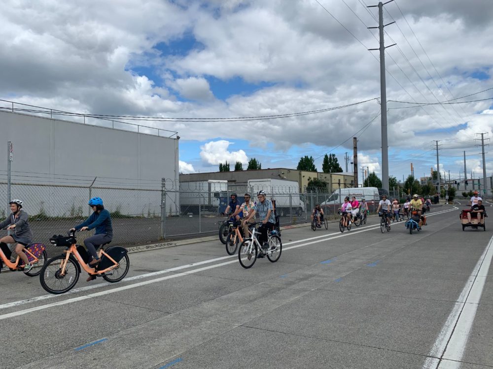 Group of people riding various bikes down empty road in industrial area.