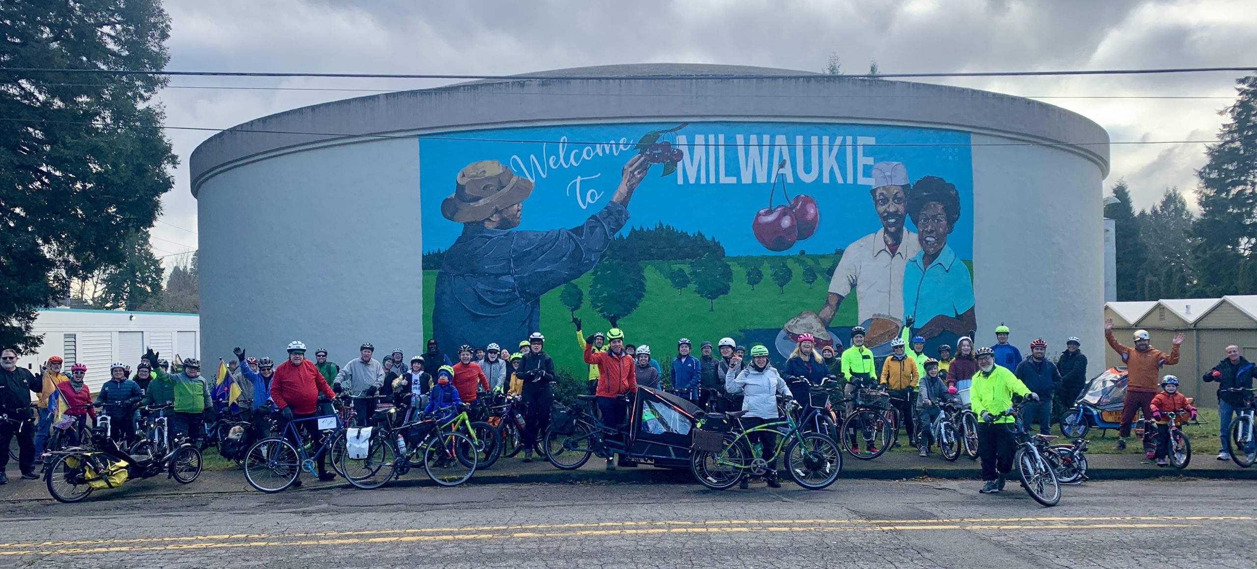 The Street Trust's 2023 New Year's Day Ride poses in front of the Welcome to Milwaukie mural by Jeremy Okai Davis featuring Ah Bing alongside Dorothy and Hurtis Hadley.