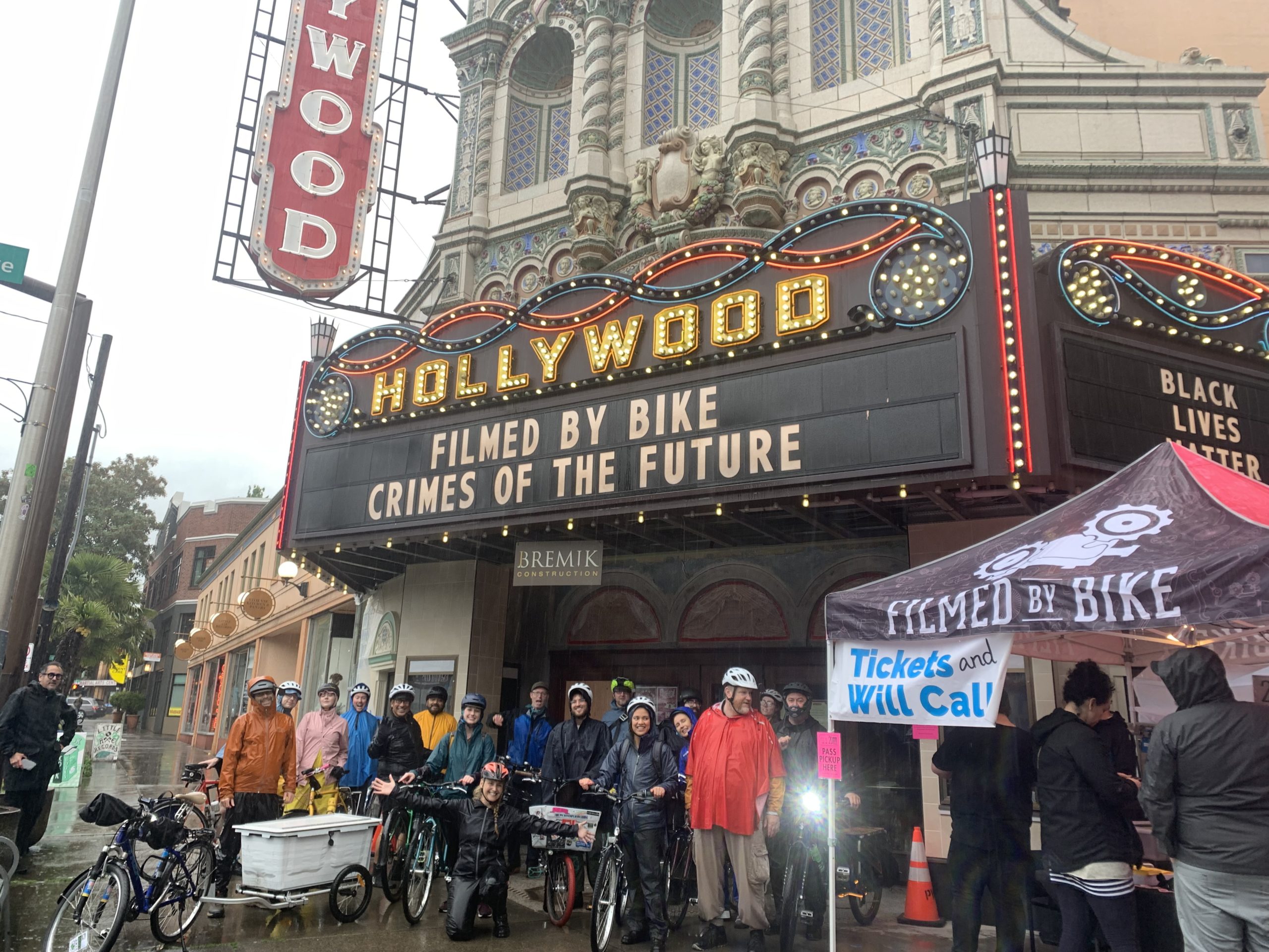 A group of people in rain gear standing under the Hollywood Theatre marquee which reads Filmed by Bike and Crimes of the Future
