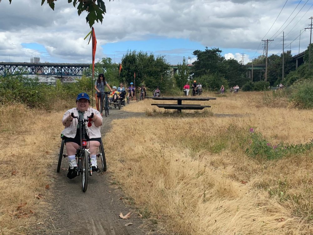 Adaptive handcycle rider rolls along gravel trail while smiling. Other riders line up on trail in background.