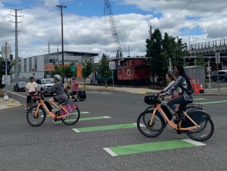 BIKETOWN e-bike riders turn left whil another cyclist waits in background and another corks traffic.