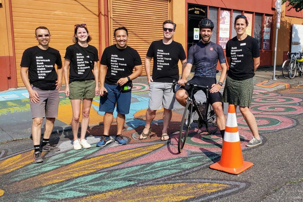 A group of six people smiling for photo on street mural outside of Milagro. One person is wearing cycling gear and sitting on their bike. Everyone else is wearing matching The Street Trust shirts.