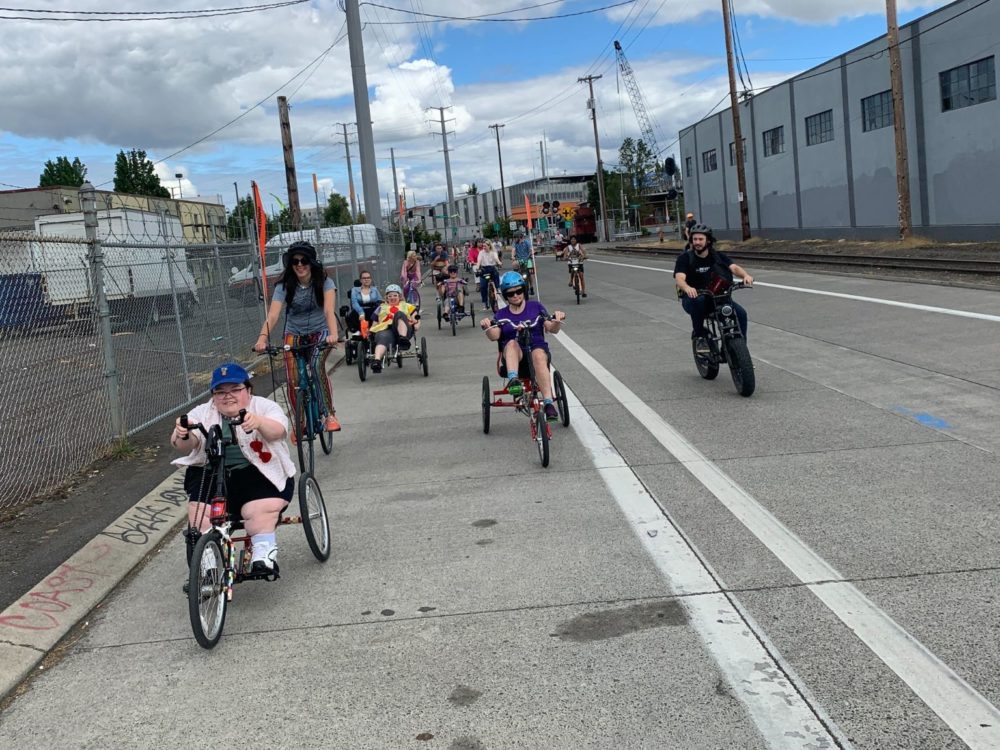A large group of people riding various adaptive and traditional bikes down a road with no traffic. Industrial buildings in background.