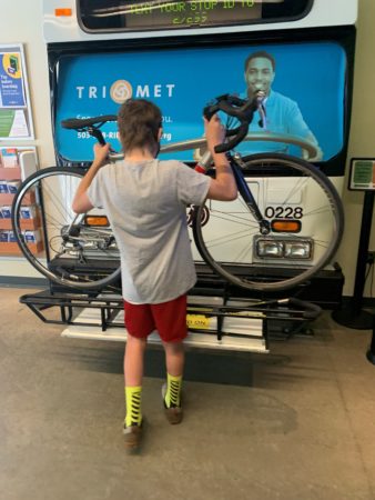 A teenager lifts a silver bike up onto a bus rack by holding its seatpost and headset