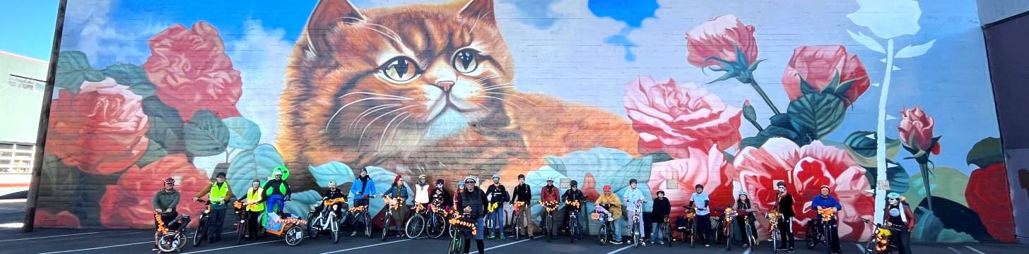 Bike riders from the 2023 Central Eastside Altares Y Muertos Bike Ride pose in front of a large mural of a kitten and roses